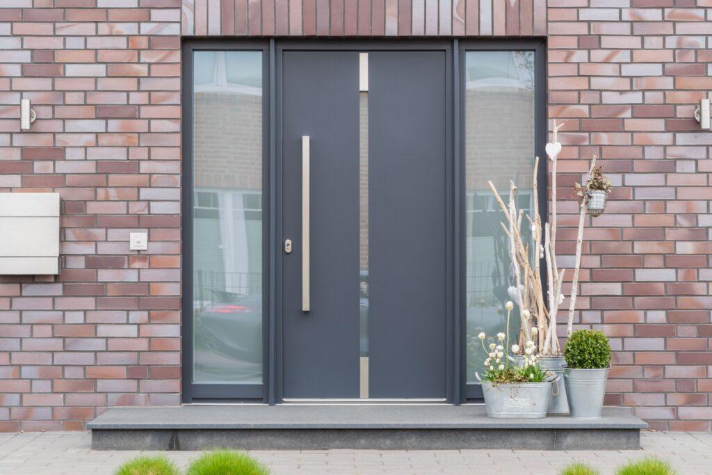 Moderne dunkelgraue Haustür mit Glaspaneelen und schlanken Metallgriffen an einem Neubau mit Ziegelsteinfassade, dekoriert mit rustikalen Elementen vor der Tür, die ein einladendes und stilvolles Erscheinungsbild bieten