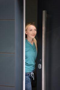 Junge Frau lächelt, während sie an der Schwelle ihrer modernen dunkelgrauen Haustür steht, die einen stilvollen und sicheren Eingang zu ihrem Zuhause bietet